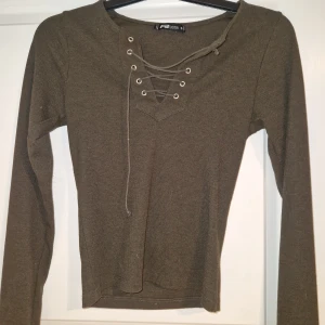 Olivgrön ribbad topp med snörning - Snygg olivgrön långärmad topp från Sister med ribbad struktur och cool snörning framtill vid halsen. Perfekt för dig som gillar en enkel men ändå trendig look. Toppen har en figurnära passform och är superenkel att styla till jeans eller kjol.