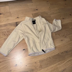 Beige fleecejacka från Everest, storlek 86/92 - En ljus fleecejacka från Everest. Jackan har en dragkedja fram och en hög krage. Perfekt för kyliga dagar. Mysig beige fleecejacka från Everest med dragkedja framtill och broderad logga på bröstet. Jackan har långärmad design och står ut med sin mjuka känsla och höga krage. Perfekt för kyliga dagar och enkel att matcha med andra plagg.