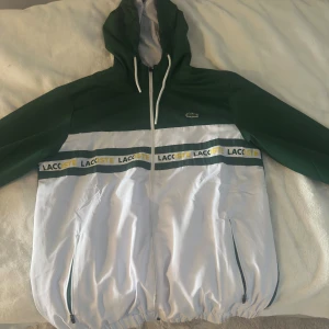 Grön och vit vindjacka från Lacoste - Snygg vindjacka från Lacoste i grönt och vitt med huva och hel dragkedja. Jackan har stora loggor framtill och baktill, samt två fickor med dragkedja. Elastisk nederkant och ärmslut. Perfekt för dig som gillar sportig och trendig stil.