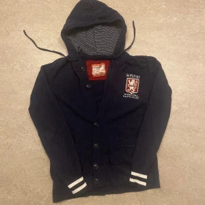 Replay cardigan med luva - Mörkblå stickad kofta från Replay i ull med huva och knappar framtill. Broderad logga och text på bröstet, stora vita siffror '1981' på ryggen. Randigt foder i huvan och vita ränder på ärmsluten ger en sportig vibe.