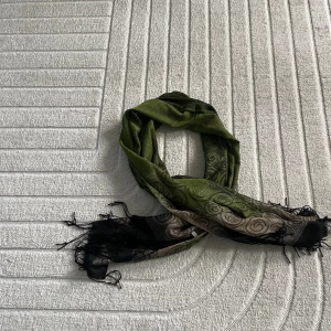 Grön och svart halsduk med spiraltryck - Snygg halsduk i grönt och svart med spiralformade mönster och fransar i ändarna. Färgerna skiftar från olivgrönt till svart och beige, vilket ger en cool och unik look. Perfekt accessoar för att lyfta din outfit.