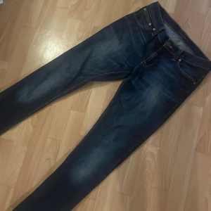 Mörkblå skinny jeans från Nudie - Snygga mörkblå skinny jeans från Nudie med klassisk femficksdesign och coola slitningar. Bakfickorna har Nudies ikoniska orangea sömmar. Jeansen är tillverkade i stretchig denim för skön passform och har normal midja. Storlek W28,L34