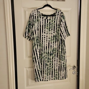 Randig kortklänning från ONLY - Snygg kortklänning från ONLY med svartvita ränder och grönt palmbladsmönster. Klänningen har rund halsringning och korta ärmar, perfekt för dig som gillar grafiska prints och tropiska vibbar. Materialet känns lätt och luftigt, vilket gör den skön att bära under varma dagar.