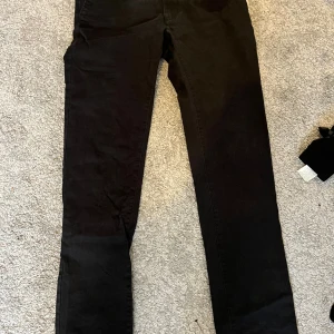 Svarta chinos Dressmann slim fit - Svarta chinos från Dressmann i slim fit-modell. Byxorna har raka ben, knappstängning bak och bälteshällor. Snyggt klassiskt snitt som passar till skjorta eller t-shirt. Materialet är mjukt och känns bekvämt mot huden.