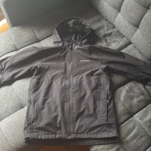 Vind och Regn jacka från Didriksons (PIKO BOYS JACKET 6) - Svart vind och  regnjacka från Didriksons med huva och hel dragkedja framtill. Jackan är tillverkad i 100% polyester och är helt vattentät. Snygg logga på bröstet och stor logotyp på ryggen. Perfekt för blöta dagar. Den är skön att använda och har justerbar luva med en metall rem innuti. Går och ta av luvan om man ej känner för att ha på den på jackan. Storlek 160. Den är använd men är fortfarande i jätte bra skick. Pris går och diskutera vid snabb affär