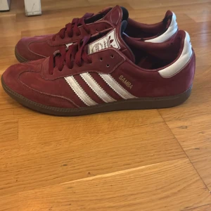 Adidas Samba vinröda sneakers - Säljer ett par Adidas Samba sneakers i vinrött med klassiska vita ränder och detaljer. Skorna har snörning, platt sula och är tillverkade i mocka och skinn. Perfekta för dig som gillar retrostil och vill sticka ut med en snygg färg på fötterna.