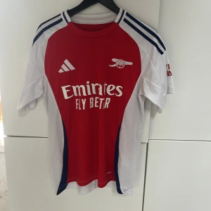 Arsenal röd och vit fotbollströja Adidas - Säljer en officiell Arsenal fotbollströja från Adidas i storlek S. Tröjan är röd med vita och blå detaljer, kortärmad och har tryck med Emirates Fly Better på bröstet samt Arsenal-loggan. .lite tecken på användning vilket syns på bilden
