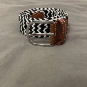 Svartvit randigt bälte med skinn - Säljer ett snyggt svartvitt randigt bälte med flätad design och bruna skinn-detaljer vid spännet. Spännet är i silverfärgad metall och bältet har vita sömmar på skinnet. Perfekt för att ge outfiten en cool touch.