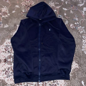 Marinblå hoodie från Polo Ralph Lauren - Säljer en marinblå hoodie från Polo Ralph Lauren med dragkedja och klassisk broderad logga på bröstet. Tröjan har huva med snörning och känguruficka framtill. Perfekt för en chill och stilren look.
