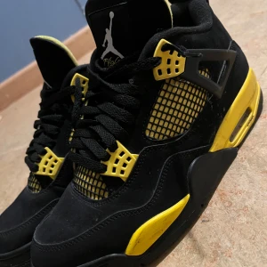 Nike Air Jordan 4 Retro svart/gul - Säljer ett par Nike Air Jordan 4 Retro i svart och gul. Skorna har klassisk meshpanel, snörning och detaljer i gult på sula och snörningsfästen. Ovandelen är i mocka och syntet, med ikonisk Jumpman-logga på plösen och sulan. Perfekta för dig som vill sticka ut med din sneakerstil.