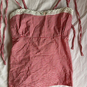 Rutig röd och vit linnetopp från Nicha Studio - Supersöt röd och vit rutig linnetopp från Nicha Studio med smala axelband, spetskant upptill och liten broderad rosdetalj framtill. Toppen har smockad rygg för extra passform och är perfekt för en somrig look.