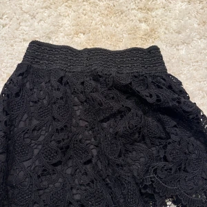 Svarta spets-shorts från Pense à toi - Säljer ett par svarta shorts från Pense à toi Paris med bred resår i midjan och snyggt virkad spets över hela plagget. De har en feminin och luftig look med fina detaljer i spetsen och lätt utställda ben. Perfekta för dig som gillar en chic och trendig stil. lite liten i storlek