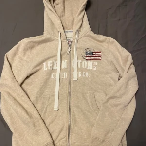 Beige hoodie från Lexington med dragkedja - Snygg beige hoodie från Lexington med dragkedja framtill, broderad amerikansk flagga på bröstet och vita snören i huvan. Klassisk college-stil med tryckt logga och kängurufickor. Perfekt för chill dagar eller när du vill ha en avslappnad look. Storlek M men sitter som S