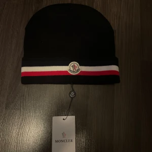 Svart Moncler mössa med ränder - Svart mössa från Moncler med klassiska ränder i blått, vitt och rött runt kanten. Mössan har Moncler-logga framtill och är ribbstickad för en snygg passform. Perfekt accessoar för att hålla stilen kallare dagar. Helt ny 