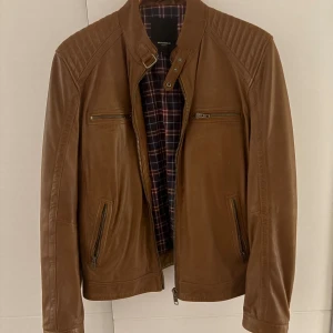 Brun skinnjacka från Rockandblue - Snygg brun skinnjacka från Rockandblue med quiltade axlar och rutigt innerfoder. Jackan har flera dragkedjor framtill, ståkrage med spänne och detaljer vid midjan. Perfekt för dig som vill ha en klassisk och stilren look.