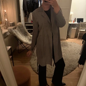 Beige/grå mocka kappa med band - Stilren trenchcoat i grå/beige mocka med klassisk krage och knytbälte i midjan. Jackan har lång ärm och rak passform. Så otroligt snygg passform och färg🩶Perfekt till höstens/vinterns middagar🥰Går inte att köpa längre så passa på🤍I jättefint skick, endast använd 1 gång av mig, skriv för fler bilder!