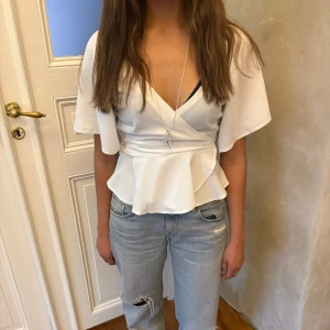 Vit omlottopp - Säljer en vit omlottopp med v-ringning, volang nedtill och knyt i ryggen. Toppen har korta, vida ärmar och en snygg figurnära passform som framhäver midjan. Materialet är mjukt och lätt, perfekt för ljusa dagar. Stilren och fräsch look!