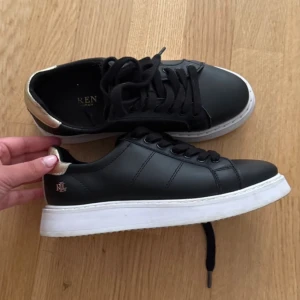 Svarta sneakers från Ralph Lauren - Snygga svarta sneakers från Lauren Ralph Lauren med vit platt sula och gulddetalj vid hälen. Skorna har snörning och RL-logga i guld på sidan. Tillverkade i skinnliknande material och har en klassisk rund tå. Perfekta för dig som gillar stilrena och trendiga sneakers.