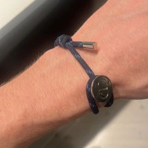 Blått Dior armband med silverdetaljer - Snyggt armband från Dior i marinblått snöre med diskreta ljusblå detaljer. Armbandet har en silverfärgad metallbricka med CD-logga och en stilren metallände. Perfekt accessoar för en trendig look.