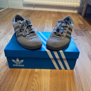 Adidas Stadt grå sneakers strl 40 - Adidas Stadt sneakers i grå mocka med klassiska tre svarta ränder på sidan och svart platt sula. Snygg retrostil med rund tå och snörning. Perfekt för dig som gillar stilrena och tidlösa sneakers med streetkänsla.