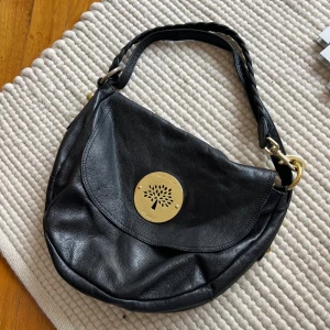 Mulberry Daria Satchel (Replika) - Snygg svart axelväska från Mulberry i mjukt skinn med flätat axelband och guldfärgade metalldetaljer. Väskan har ett stort Mulberry-emblem framtill och innerfoder med Mulberry-mönster samt dragkedja. Perfekt storlek för det viktigaste. Jag är osäker på väskans äkthet därav det låga priset, en garanterad äkta går för flera tusen. Det finns slitage på väskan där innertyget har släppt i vissa sömmar, även en av guldringarna på sidan har tappat lite av sin guldiga färg.