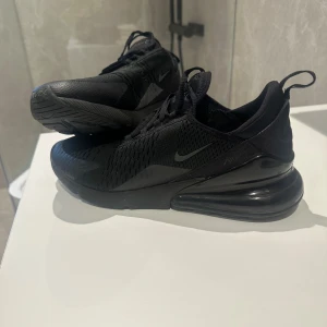 Nike Air Max 270 helsvarta sneakers - Säljer ett par helsvarta Nike Air Max 270 sneakers med snygg mesh-överdel och stor synlig Air-enhet i hälen. Skorna har snörning, rund tå och platt sula. Perfekta för dig som gillar sportig och clean stil. Materialet är syntet och textil.
