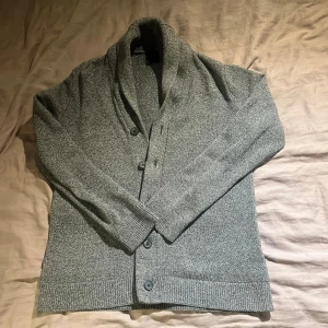 Grå kofta med sjalkrage från H&M - Snygg gråmelerad kofta från H&M med sjalkrage och knappar framtill. Stickad i mjukt bomullsmaterial, perfekt för lager-på-lager. Långärmad och med ribbade muddar vid ärmslut och nederkant. Enkel att matcha med jeans eller chinos.