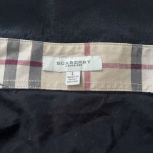 Svart Burberry skjorta med rutig krage - Svart skjorta från Burberry i storlek s med klassiska rutiga detaljer på krage och manschetter. Skjortan har knappar framtill och Burberrys logga broderad på bröstet. Materialet är mjuk bomull och passformen är normal. Perfekt för dig som gillar stilrena och ikoniska plagg.