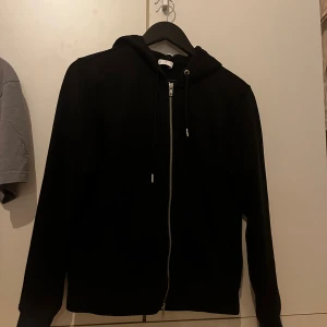 Svart zip hoodie från Asket  - Svart hoodie från Asket i storlek XS, har krympt i tvätten så passar 15 år, med hel dragkedja framtill och snörning vid huvan. Tröjan har lång ärm och klassisk passform, perfekt för chill dagar. Materialet är mjuk bomull och insidan är skönt borstad. Snygg och enkel design utan tryck.
