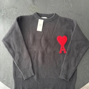 Svart stickad tröja från Ami - Mysig grå stickad tröja från Ami med rund hals och ribbade muddar. På bröstet sitter en stor röd hjärtapplikation med bokstaven A. Perfekt för dig som vill ha en stilren men ändå unik look.