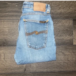 Nudie jeans - Säljer ett par ljusblå slim jeans från Nudie Jeans med klassiska orange sömmar och snygg broderad bakficka. Modellen har normal passform och raka ben. Midja 33cm, längd 94cm. Modellnamn okänt.  Jeansen kan möjligtvis vara uppsydda då dem är korta för att vara L32 så kolla måtten noggrant :) Ifall det inte finns någon bild i annonsen där jag bär plaggen jag säljer så är det för att jag ej kan ha storleken heller passformen, så kolla måtten för storlek och googla på modellnamnet för passform 😉