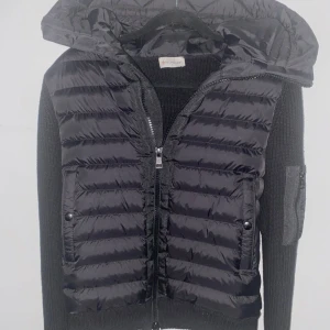 Svart Moncler pufferjacka med huva - Svart pufferjacka från Moncler med stickade ärmar och vadderad kropp. Jackan har huva, två fickor med knappstängning och dragkedja framtill. Snyggt quiltad design och Moncler-logga på ärmen. Perfekt för dig som gillar exklusiva streetwear-vibbar.