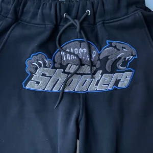 Svarta Trapstar mjukisbyxor med broderi - Svarta mjukisbyxor från Trapstar med stort broderat Shooters-motiv i grått och blått framtill. Byxorna har snörning i midjan och dragkedja vid benslut. Sköna och stilrena, perfekta för chill eller streetwear.