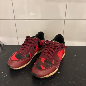 Valentino Garavani Rockrunner sneakers - Snygga Valentino Garavani Rockrunner sneakers i rött och vinrött med coolt kamouflagemönster. Skorna har detaljer i mocka och skinn, svarta och röda nitar på hälen samt klassisk snörning. Perfekta för dig som vill sticka ut med exklusiva och trendiga sneakers.