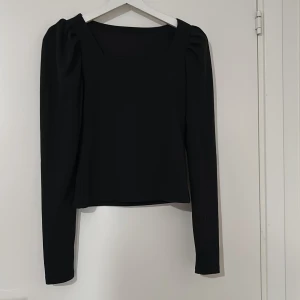 Svart långärmad blus med puffärm - Säljer en svart blus med lång ärm och snygg fyrkantig halsringning. Toppen har puffade axlar som ger en cool siluett och är croppad i modellen. Materialet känns mjukt och har lite stretch, perfekt för att styla med jeans eller kjol.