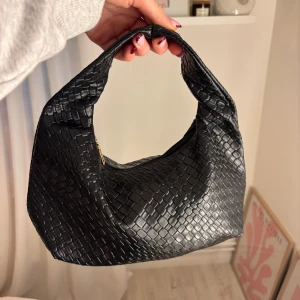 Svart oanvänd handväska - Snygg svart oanvänd handväska med reptilinspirerat mönster och den har en dragkedja. Väskan har en mjuk, rundad form och är tillverkad i skinnimitation. Perfekt storlek för att få plats med det viktigaste! Aldrig använd! 🖤