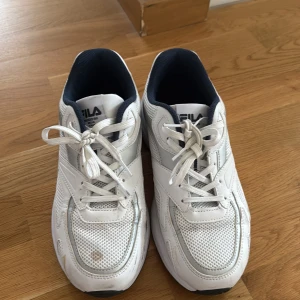 Vita sneakers från FILA - Säljer ett par klassiska vita sneakers från FILA med mesh och syntetdetaljer. Skorna har snörning, rund tå och en chunky sula. Insidan är fodrad i mörkblått tyg och loggan syns tydligt på både plös och häl. Perfekta för en sportig och clean look.