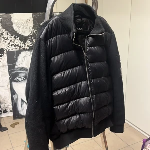 Svart pufferjacka från BVALDI - Svart pufferjacka från BVALDI med stickade ärmar och rygg. Jackan har quiltad framsida i glansig polyester, ribbad krage och muddar samt dragkedja framtill. Snygg patch på ena ärmen, storlek S.