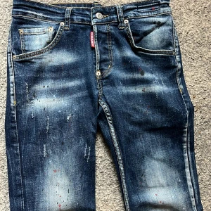 Dsquared2 blå jeans - Snygga blå jeans från Dsquared2 i storlek 48 med coola slitningar och färgstänk för en edgy vibe. Klassisk femficksmodell med Dsquared2-logga på bakfickan. Jeansen har en rak passform och är tillverkade i robust denim.