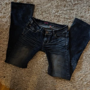 Mörkblå lågmidjade/utsvängda jeans med kedjedetaljer - Snygga mörkblå jeans från JNY Basic med slitningar och coola kedjedetaljer på bakfickorna. Klassisk femficksmodell med kontrastsömmar och rak passform. Perfekt för dig som gillar en lite edgy stil och vill sticka ut med detaljer.