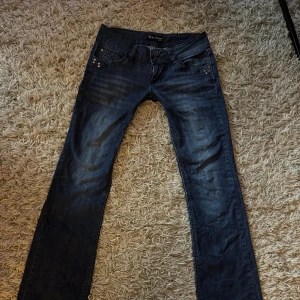 Mörkblå lågmidjade /utsvängda jeans - Snygga mörkblå jeans från Miss Olive med raka ben och coola detaljer som nitar på bakfickorna. Jeansen har klassisk femficksdesign, kontrastsömmar och låg midja. Perfekt för dig som gillar en stilren look med lite edge.