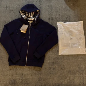 Marinblå Burberry hoodie med dragkedja - Snygg marinblå hoodie från Burberry med klassiskt rutigt foder i huvan. Tröjan har dragkedja framtill, snörning i huvan och ribbade muddar. Perfekt för dig som vill ha en stilren och exklusiv look med ikoniska Burberry-detaljer.