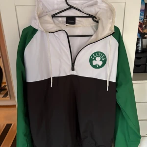 Boston Celtics vindjacka från New Era - Snygg Boston Celtics vindjacka från New Era i grönt, vitt och svart. Jackan har huva med snörning, meshfoder och Celtics-logga på bröstet. Half zip framtill och elastisk nederkant. Perfekt för dig som gillar basket och streetwear.