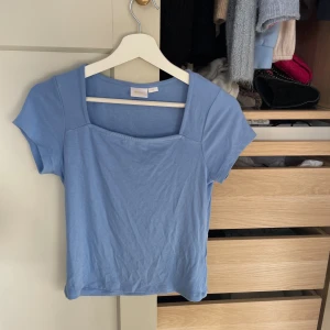 Ljusblå topp med fyrkantig ringning - Säljer en ljusblå topp med korta ärmar och snygg fyrkantig ringning. Toppen är i mjuk bomull och har en enkel, stilren design som passar perfekt till jeans eller kjol. Perfekt för vår och sommar!