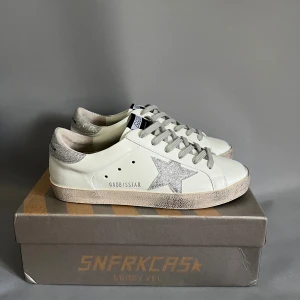 Golden Goose - Golden Goose Superstar sneakers i vitt skinn med silverglittrig stjärna på sidan och silverdetaljer på hälen. Grå snörning och platt sula. Klassisk låg modell med svart GGDB-märke på plösen. Perfekta för dig som gillar en clean men ändå glittrig look. Storlek 39 passar även 40🤝 använda 1 gång