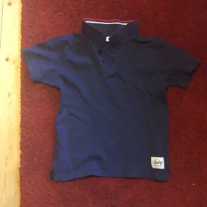 Mörkblå Andy by Frank Dandy piké - Snygg mörkblå pikétröja från Andy by Frank Dandy med klassisk krage och korta ärmar. Tröjan har en liten etikett nertill och diskreta knappar vid halsen. Perfekt för en stilren och avslappnad look.