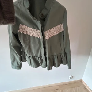 Grön jacka med spetsdetaljer - Snygg grön jacka med volangkant nertill och dekorativa spetsband med små bollar över bröstet och ryggen. Jackan har knappar framtill och är långärmad. Köpt begangnad, men aldrig använd själv. Men bra skick! 