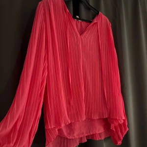 Rosa blus med v-ringning - Säljer en snygg blus i stark rosa färg med v-ringning och långa, luftiga ärmar. Blusen har tunna vertikala ränder och ett flowigt, lite längre bakstycke. Perfekt för dig som gillar färg och volym i din outfit.