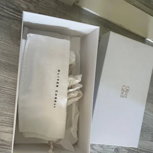 Oliver Cabell vita sneakers med svart detalj - Säljer ett par vita sneakers från Oliver Cabell med svart häl och grå cirkeldetalj på sidan. Skorna har snörning och är tillverkade i skinn med platt sula. Kommer i originalkartong och dustbag. Perfekta för dig som gillar stilrena och trendiga sneakers.