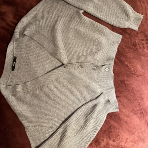 Grå ribbad kofta från Bik Bok - Grå ribbstickad kofta från Bik Bok med v-ringning och knappar framtill. Modellen har långa ärmar och breda muddar vid ärmslut och nederkant. Perfekt att slänga över en t-shirt eller linne för en chill look. Har ett litet hål vid högra axeln. Köparen står för frakten